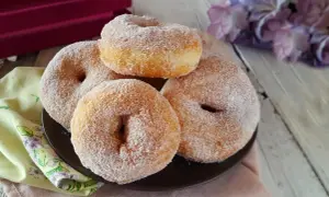 Graffe senza patate, soffici e dorate, ricoperte di zucchero. Un dolce invitante su un piatto scuro, con fiori viola sullo sfondo.