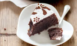 Una fetta di torta caprese ricetta facile, cosparsa di zucchero a velo e cacao, su un piatto bianco a forma di cuore. Un cucchiaino ha prelevato un pezzetto.