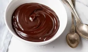 Una ciotola bianca piena di glassa al cioccolato veloce, lucida e invitante, con due cucchiai d'argento accanto.