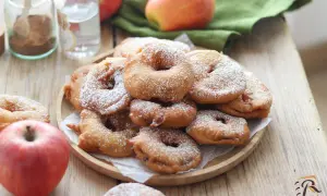 Frittelle di mele della nonna, dorate e cosparse di zucchero a velo, su un piatto di legno, con mele fresche e spezie sullo sfondo.