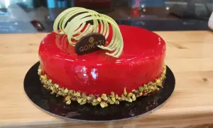 Torta con glassa a specchio lamponi, decorata con riccioli di cioccolato bianco e pistacchi tritati.