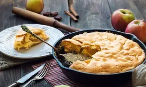 Una deliziosa torta di mele in padella, appena sfornata, con una fetta servita su un piatto. Mele fresche e cannella completano la scena rustica.
