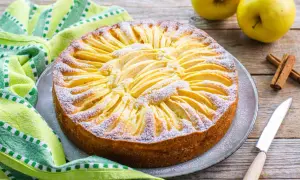 Torta di mele con yogurt, decorata con fettine di mela e zucchero a velo, su un piatto grigio, accanto a un tovagliolo verde e bastoncini di cannella.