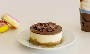 Mini cheesecake al cioccolato con base croccante e copertura di cioccolato fondente, accompagnato da due ciambelle colorate e una tazza sorridente.