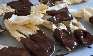 Chiacchiere al cioccolato, dolci croccanti e friabili, cosparse di zucchero a velo e immerse in golosa cioccolata fondente.