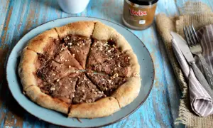 Pizza al cioccolato con crema Twix e nocciole, cosparsa di granella croccante. Un dolce goloso pronto da gustare.