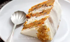Una fetta di torta di carote, soffice e profumata, con glassa bianca e noci tritate. Accanto, un cucchiaino d'argento.