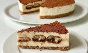 Una fetta di cheesecake tiramisù, con savoiardi inzuppati e cacao in polvere, su un piatto bianco.