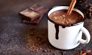 Una tazza di cioccolata calda fondente fumante, con stecca di cannella e cacao sparso. Pezzi di cioccolato fondente sullo sfondo.