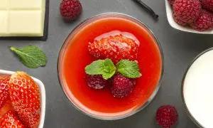 Un bicchiere di coulis di fragole, guarnito con fragola, lamponi e menta. Accanto, fragole fresche, cioccolato bianco, latte e bacca di vaniglia.