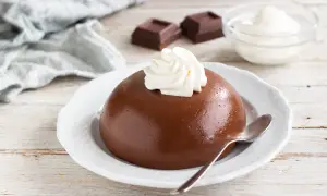 Budino al cacao liscio e lucido, guarnito con panna montata, su un piatto bianco. Accanto, cubetti di cioccolato e una ciotola di panna.