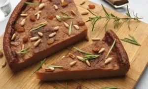 Due fette di castagnaccio morbido, guarnite con pinoli, uvetta e rosmarino, su un tagliere di legno.