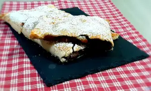 Strudel mele e cioccolato cosparso di zucchero a velo, su una pietra nera e tovaglia a quadri rossi.