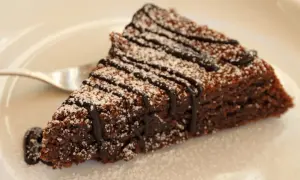 Una fetta di torta caprese al cioccolato, cosparsa di zucchero a velo e con una colatura di cioccolato fondente.