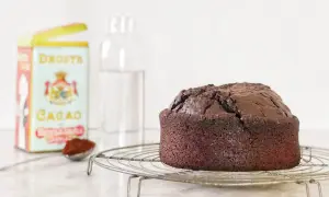 Torta all'acqua al cacao appena sfornata, su una gratella. Accanto, una scatola di cacao Droste e un bicchiere d'acqua.