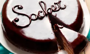 Una lucida torta viennese al cioccolato, con una fetta tagliata, decorata con la scritta "Sacher" in glassa scura.