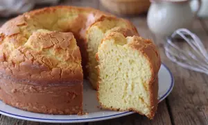 Una torta alta, soffice e dorata, con una fetta tagliata che rivela la sua morbidezza. Sullo sfondo, uova e latte, ingredienti per un dolce fatto in casa.