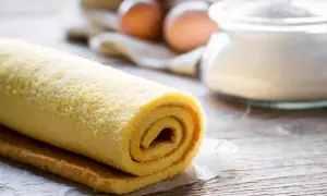 Rotolo di pasta biscotto soffice, pronto per essere farcito. Sullo sfondo, uova e zucchero, ingredienti essenziali per un dolce fatto in casa.