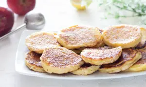Piatto di frittelle di mele dorate e soffici, spolverate di zucchero a velo. Sullo sfondo, mele rosse e un colino.