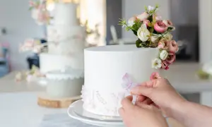 Una mano aggiunge una farfalla a una torta bianca decorata con fiori e paillettes. Sullo sfondo, un'altra torta a più piani con decorazioni floreali rosa.