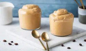 Due bicchieri di cremosa schiuma di caffè, perfetti per una ricetta crema di caffè senza panna. Due cucchiaini dorati e chicchi di caffè completano la scena.