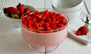 Delizioso mousse di fragole in una coppa di vetro, guarnito con fragole fresche a pezzetti. Accanto, una ciotola di fragole e ciotole bianche.