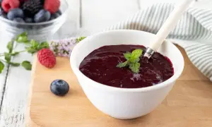 Una ciotola bianca contiene un denso coulis frutti di bosco, guarnito con menta fresca. Frutti di bosco freschi e foglie di menta sono sparsi sul tagliere.
