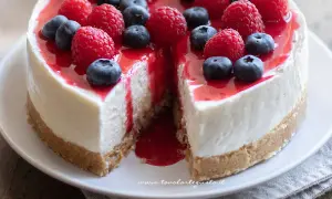 Cheesecake ricetta originale con frutti di bosco freschi e coulis rosso. Una fetta è stata tolta, rivelando la sua consistenza cremosa.