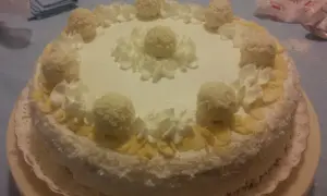 Torta Raffaello ricoperta di cocco, decorata con panna montata e praline, un'esplosione di dolcezza.
