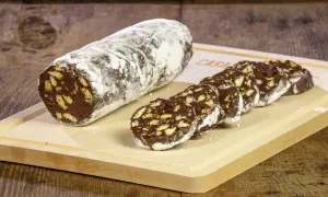 Un rotolo di salame al cioccolato, cosparso di zucchero a velo, è affettato su un tagliere di legno. Pezzi croccanti di biscotto si mescolano al cioccolato fondente.