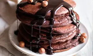Pancake cacao golosi, impilati e ricoperti da una cascata di salsa al cioccolato fondente e nocciole intere.