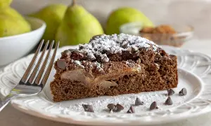 Una fetta di torta pere e gocce di cioccolato, cosparsa di zucchero a velo e gocce di cioccolato, su un piatto bianco con una forchetta.