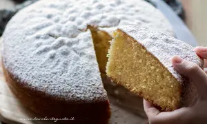 Una fetta di soffice torta margherita, cosparsa di zucchero a velo, viene sollevata da una mano.