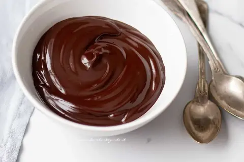 Una ciotola bianca piena di glassa al cioccolato veloce, lucida e invitante, con due cucchiai d'argento accanto.