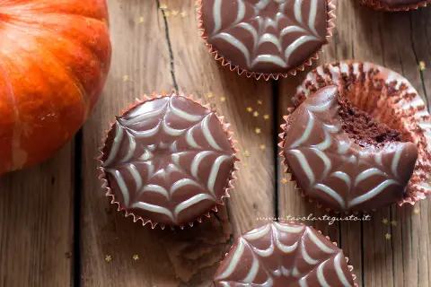 Muffin Halloween facili con decorazione a ragnatela. Una zucca arancione e stelline dorate completano la scena.