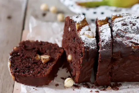 Fetta di banana bread cacao con nocciole e zucchero a velo, su tagliere di legno.