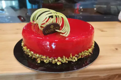 Torta con glassa a specchio lamponi, decorata con riccioli di cioccolato bianco e pistacchi tritati.