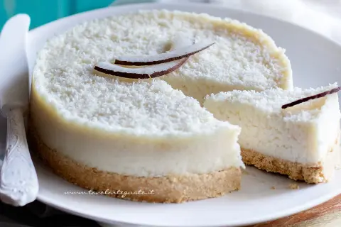 Delizioso cheesecake al cocco, ricoperto di cocco grattugiato e decorato con scaglie di cocco fresco. Una fetta è già stata tagliata.