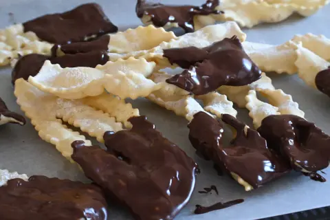Chiacchiere al cioccolato, dolci croccanti e friabili, cosparse di zucchero a velo e immerse in golosa cioccolata fondente.