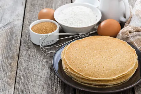 Ingredienti per la ricetta crepes: farina, uova, zucchero, latte e frusta su un tavolo di legno.