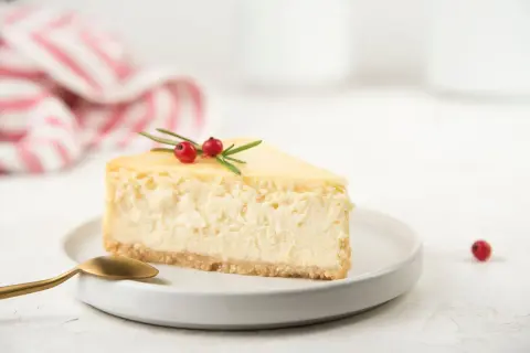 Una fetta di cheesecake con yogurt greco, guarnita con bacche rosse e rosmarino, su un piatto bianco con un cucchiaio dorato.