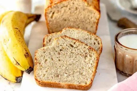 Fetta di banana bread senza uova, soffice e dorata, accanto a banane mature e un bicchiere di latte.