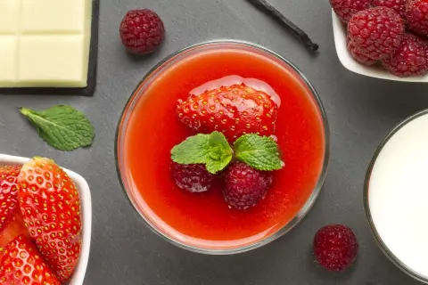 Un bicchiere di coulis di fragole, guarnito con fragola, lamponi e menta. Accanto, fragole fresche, cioccolato bianco, latte e bacca di vaniglia.