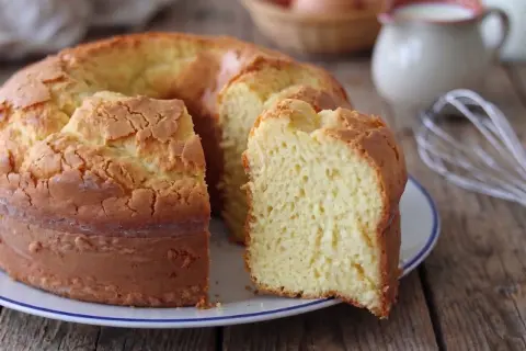 Una torta alta, soffice e dorata, con una fetta tagliata che rivela la sua morbidezza. Sullo sfondo, uova e latte, ingredienti per un dolce fatto in casa.