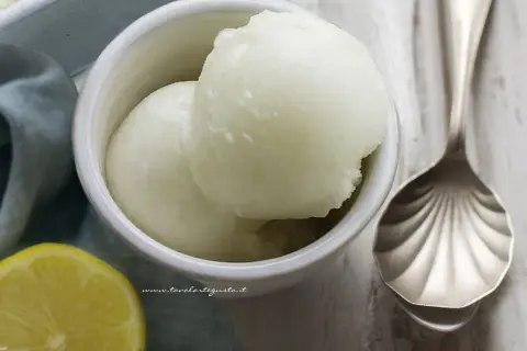 Coppetta con due palline di sorbetto al limone alcolico, accanto una fetta di limone e un cucchiaio decorato.