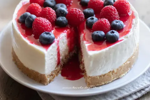 Cheesecake ricetta originale con frutti di bosco freschi e coulis rosso. Una fetta è stata tolta, rivelando la sua consistenza cremosa.