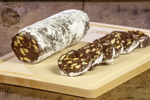Un rotolo di salame al cioccolato, cosparso di zucchero a velo, è affettato su un tagliere di legno. Pezzi croccanti di biscotto si mescolano al cioccolato fondente.