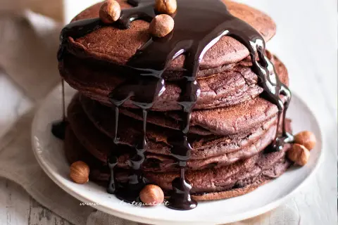 Pancake cacao golosi, impilati e ricoperti da una cascata di salsa al cioccolato fondente e nocciole intere.
