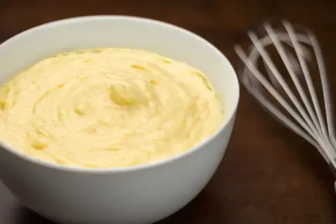 Crema pasticcera senza glutine, liscia e vellutata, pronta per farcire dolci. Vicino, una frusta in movimento.
