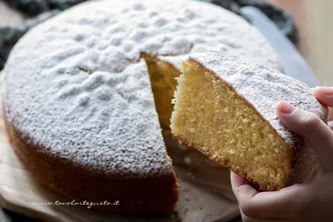 Una fetta di soffice torta margherita, cosparsa di zucchero a velo, viene sollevata da una mano.